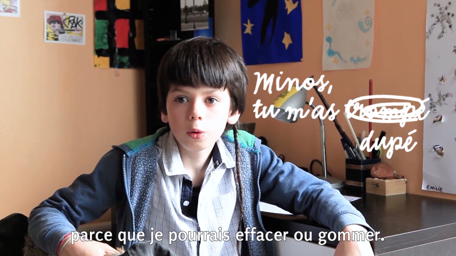 Capture d'écran d'une interview d'un élève, en plan poitrine, assis dos à son bureau, dans sa chambre. L'intervention typographique manuscrite dit : "Minos, tu m'as trompé dupé", mais le mot "trompé" est barré.