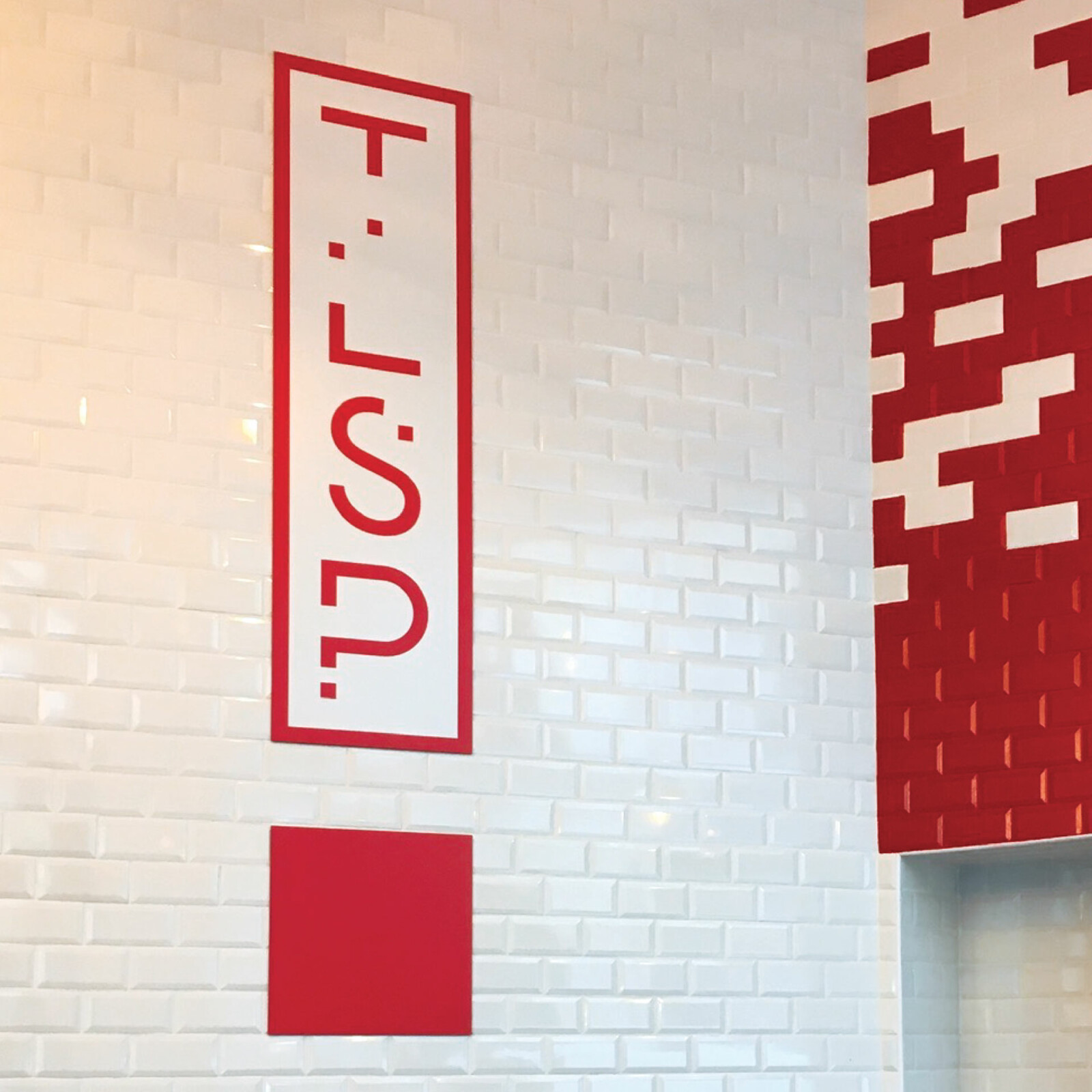 Photo du logo TLSP imprimé sur Dibond, collé au mur, dans un rectangle blanc, un carré rouge sous celui-ci crée un point d’exclamation, le carrelage rouge et blanc est visible.