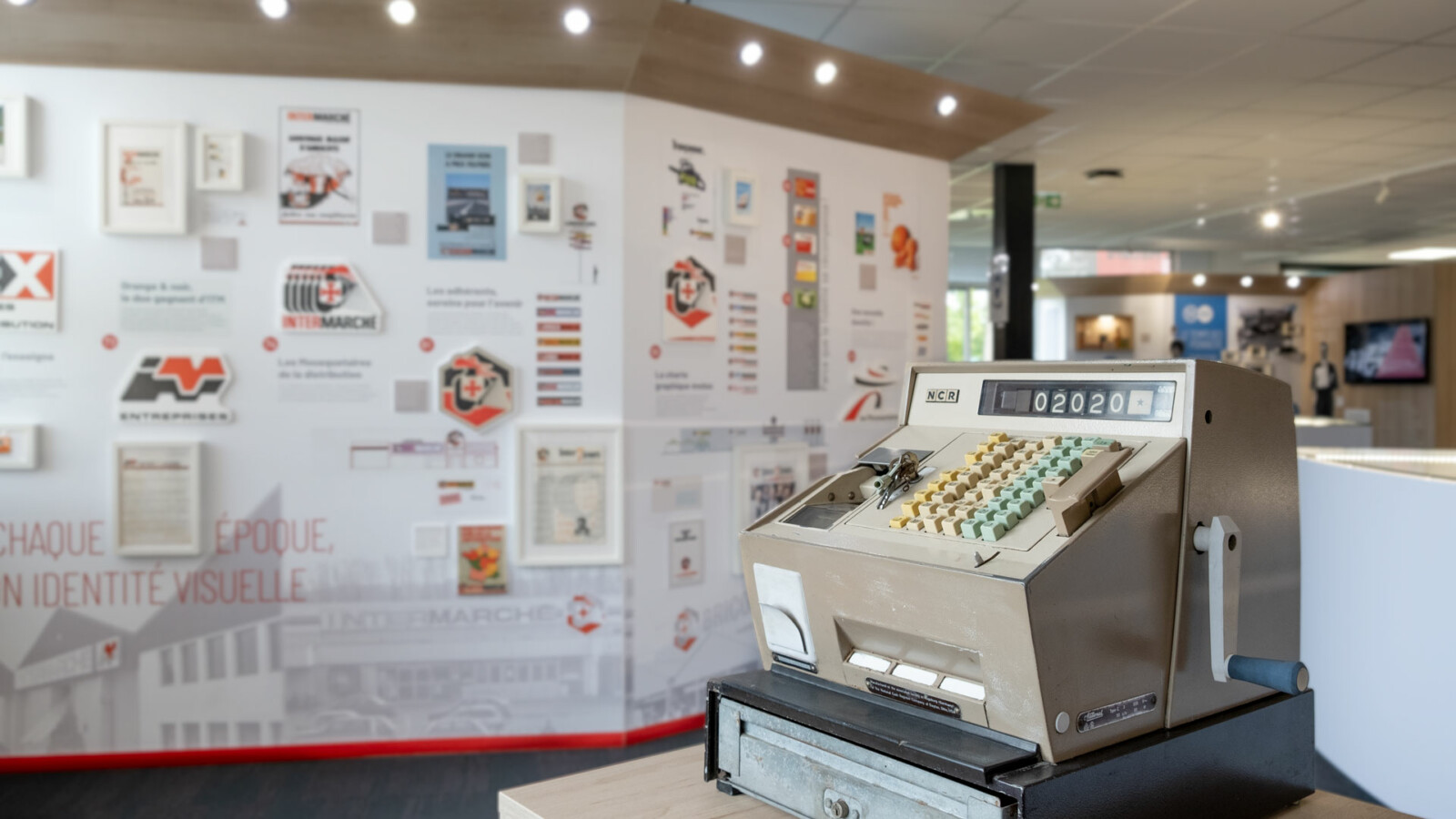 Une caisse enregistreuse des années 1970 est exposée au sein du musée d’entreprise des Mousquetaires, devant les panneaux racontant l’histoire des logos des enseignes du Groupement.