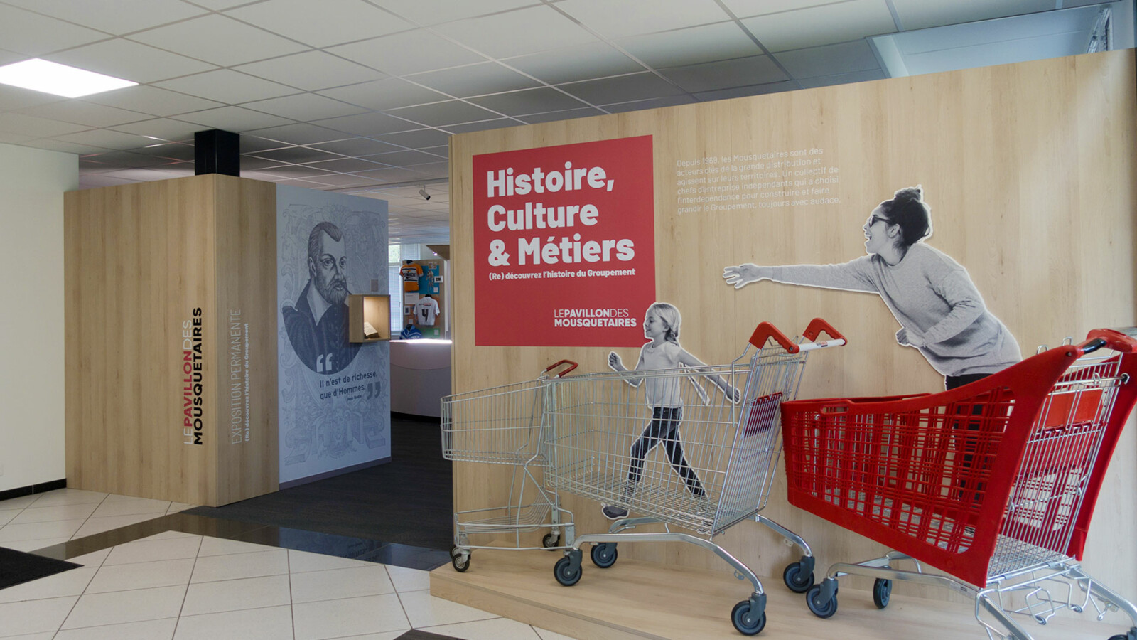 Vue de l’entrée du musée d’entreprise des Mousquetaires. Trois caddies de supermarché sont exposés sous le panneau rouge « Histoire, Culture & Métiers -Le Pavillon des Mousquetaires ».
