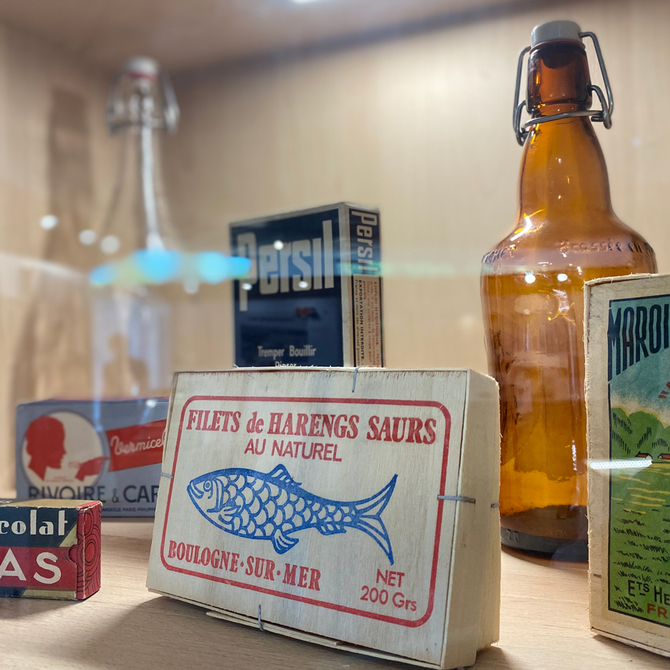 Vitrine de produits alimentaires et ménagers des années 1980 : une boîte de filets de harengs saurs, une bouteille de bière, une boîte de lessive Persil…