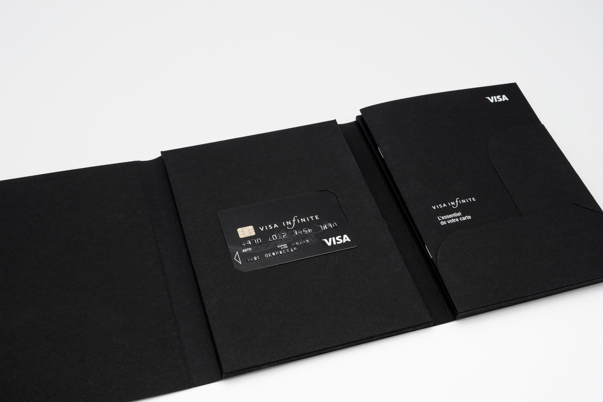 Vue du Welcome Pack ouvert, deux plis roulés, sur la droite dans un rabat se trouve le guide d’utilisation, au centre dans deux encoches la carte Visa Infinite l'ensemble est constitué de papier noir teinté masse.
